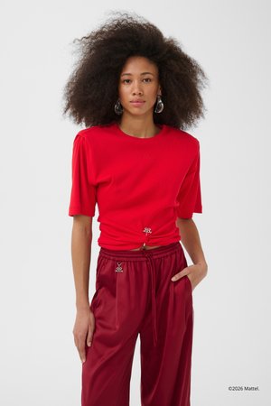 Kvinde med fyldigt, krøllet hår iført rød, samlet top med korte ærmer og matchende satinbukser med hånden i lommen mod hvid baggrund.