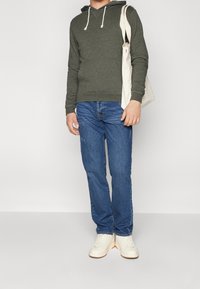 Persona che indossa una felpa verde oliva, jeans blu, sneaker bianche e porta una borsa a tracolla beige su una spalla, su uno sfondo neutro.