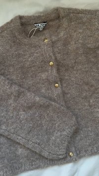 Cardigan tricoté marron clair avec col rond, bords côtelés et quatre boutons décoratifs dorés sur le devant. Tissu doux et texturé.