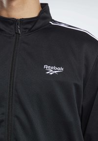 Černá běžecká bunda Reebok se stojáčkovým límcem, zapínáním na zip a bílými akcentními pruhy na ramenou s vyšitým logem na hrudi.