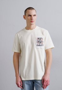 Kreemi värvi puuvillane T-särk, millel on tasku, kus on trükitud oranžide ja hallide hoonete muster. Relaxeeritud lõige ja lühikesed varrukad.