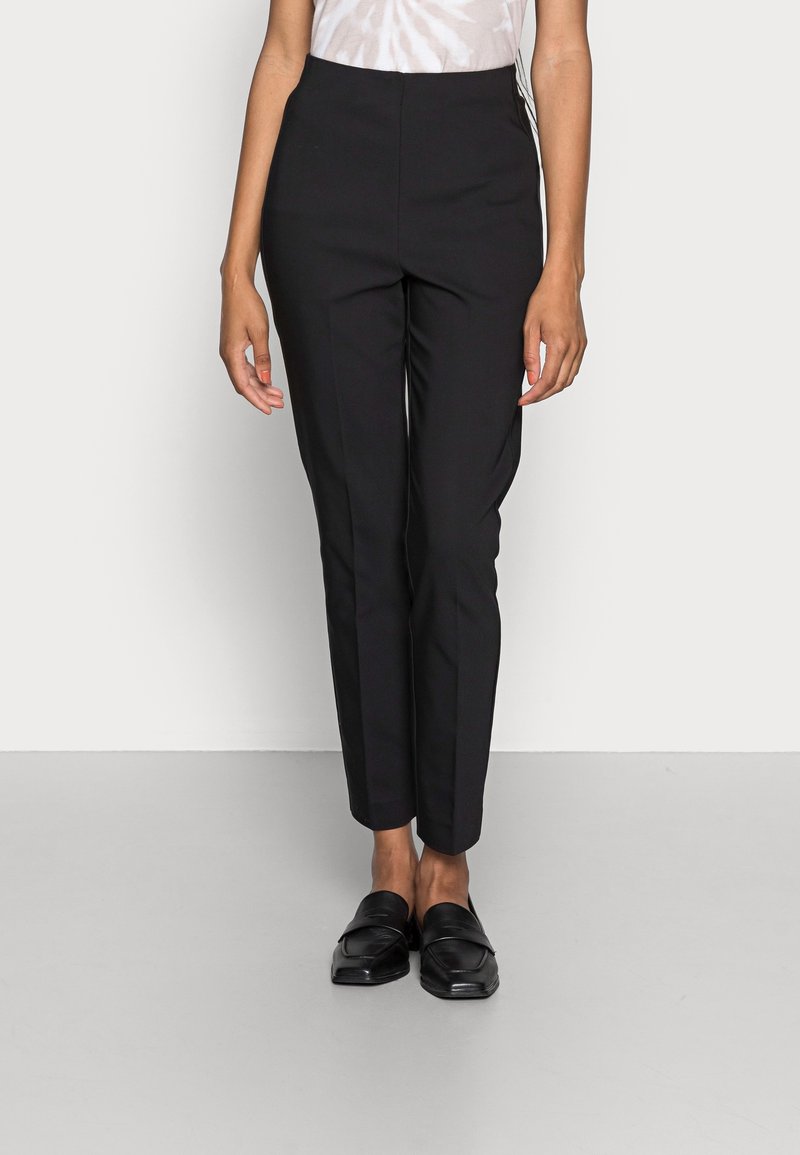 Woman wearing fitted black dress pants and black loafers, standing against a plain light background.