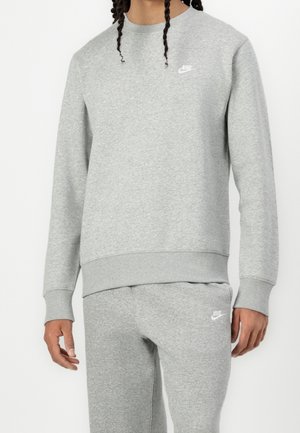 Persoon draagt een lichtgrijze Nike-trui en bijpassende joggingbroek met witte Nike-logo's op de borst en dij.