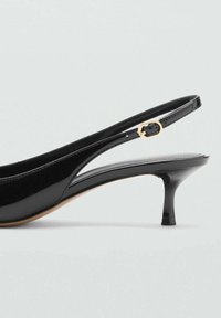 Escarpin en cuir verni noir avec un bout pointu, une fine bride pour la cheville et un petit talon effilé. Accent de quincaillerie dorée sur la bride.