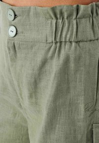Pantalones de lino verde claro que cuentan con dos botones frontales, una cinturilla fruncida y detalles de costura sutiles. La tela tiene un acabado texturizado.
