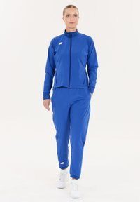 Blauer Sport-Jogginganzug aus glattem, leichtem Stoff. Besitzt eine Jacke mit Reißverschluss und taillierte Hosen mit einem Logo auf der Brust und dem Bein.