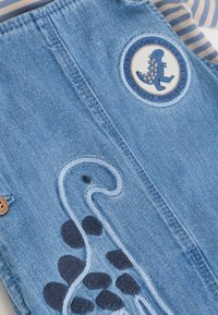 Prenda de denim con parche de dinosaurio y diseño de dinosaurio bordado, llevada sobre un fondo de tela a rayas en tonos beige y azul.