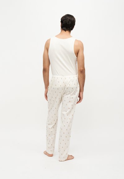 Homme debout pieds nus, de dos, portant un débardeur blanc sans manches et un pantalon de pyjama blanc avec de petits motifs imprimés.