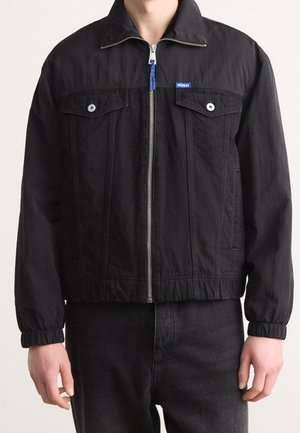 Homme portant une veste noire zippée avec des poches poitrine à boutons et des poignets élastiques, associée à un pantalon noir, visible du cou aux hanches.