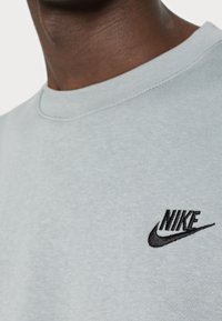 Ljusgrå sweatshirt med ribbad halsringning, med en svart broderad Nike-logotyp på vänster sida. Slät textur.