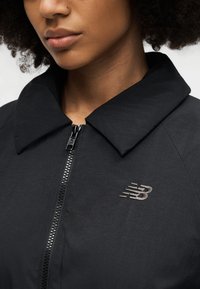 Κοντινό πλάνο προσώπου ατόμου που φοράει μαύρο μπουφάν με φερμουάρ και φαρδιά γιακά, με το λογότυπο New Balance στο στήθος, εστίαση στο κάτω μέρος του προσώπου και στον λαιμό.