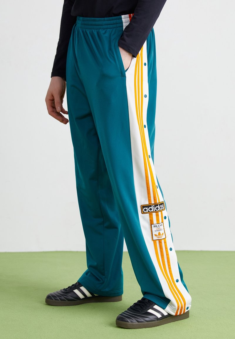 Tealfärgade träningsbyxor med vita sidosträck som har tre orange linjer och tryckknappar. Inkluderar en Adidas-logotyp. Lättviktsmaterial.