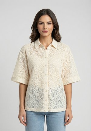 Femme portant une chemise boutonnée à manches courtes en dentelle crème avec col, associée à un jean bleu clair, debout devant un fond uni.