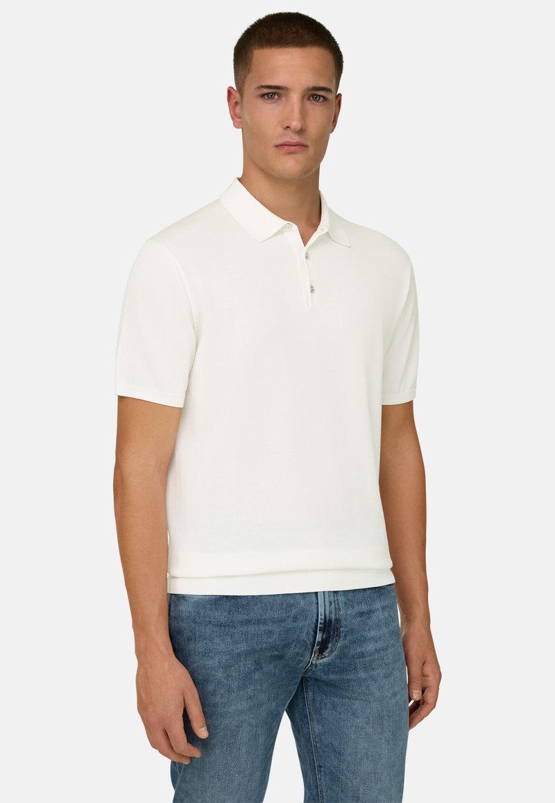 Homme portant un polo à manches courtes blanc et un jean bleu, debout devant un fond clair uni, faisant face à l'avant.