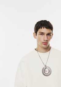 Mladý muž s krátkými tmavými vlasy, oblečený ve světlém svetr s kapucí a velkým kulatým stříbrným náhrdelníkem s obrázkem psa.