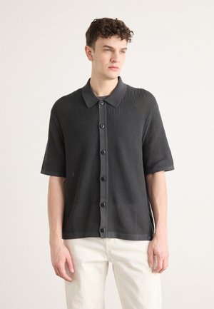 Short-sleeve, dark gray knit polo shirt with a button-up front, collar, and ribbed texture. Paired with light-colored pants.