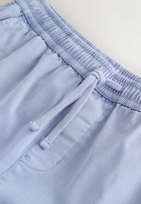 Pantalon à taille élastique bleu clair avec cordon de serrage noué et coutures visibles sur la poche latérale, en tissu lisse.