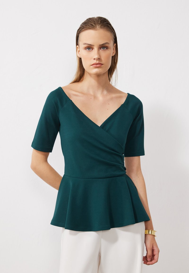 Teal peplum topp med buet V-hals og korte ermer. Glatt stoff med lett stretch og vid hem, brukt med hvite bukser.