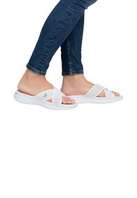 White slide sandals with perforated cross straps, textured sole, and a lightweight design, paired with blue denim skinny jeans.