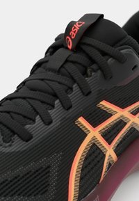 Chaussure de sport noire avec un dessus texturé, des détails rayés orange et noir, et une semelle rose. Logo Asics affiché en évidence sur la languette.