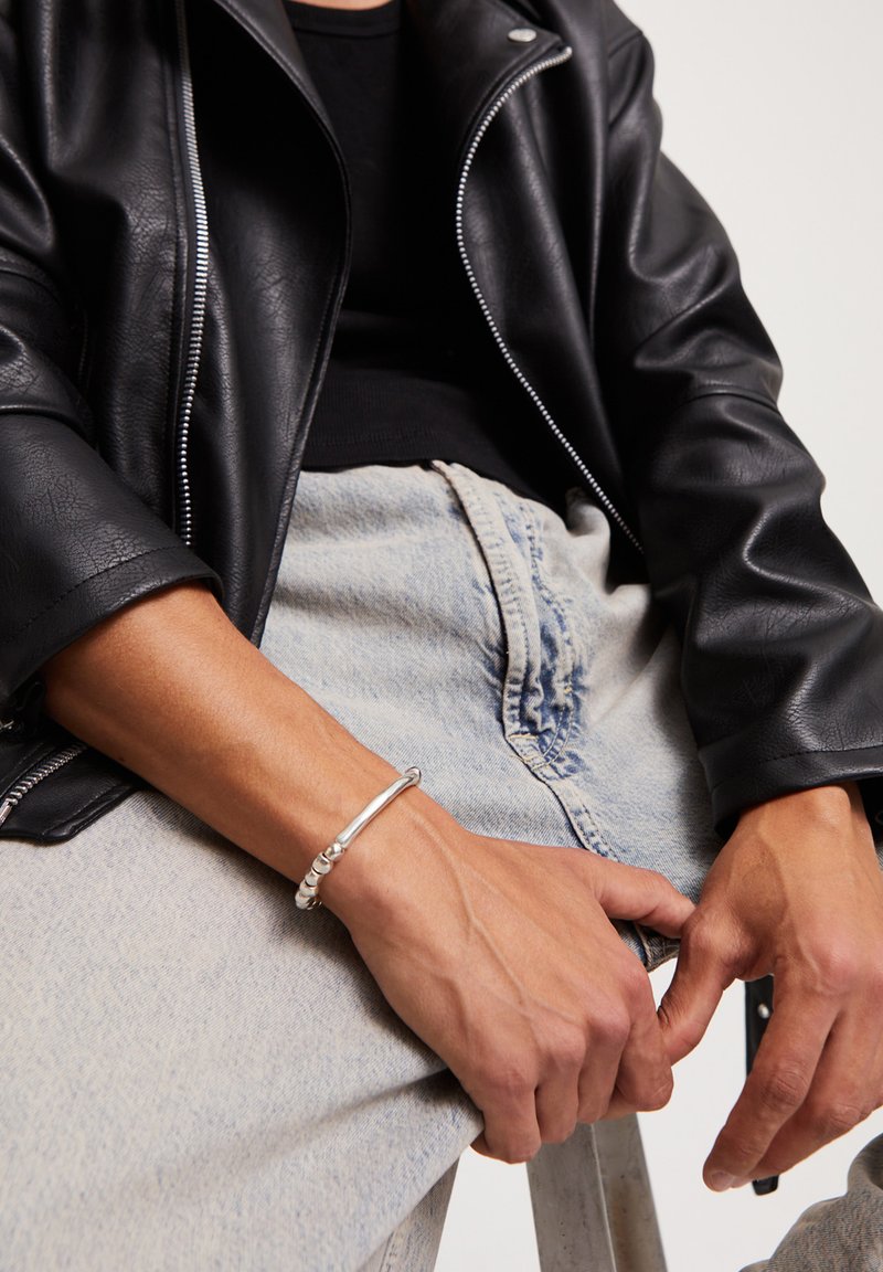 Persona vistiendo jeans azul claro, chaqueta de cuero negra y pulsera plateada, sentada con las manos descansando sobre las piernas.