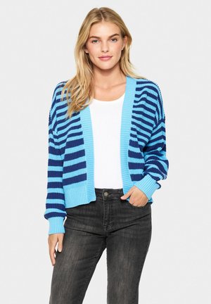Cardigan rayé bleu et marine avec des poignets et un ourlet côtelés. Porté par-dessus un haut blanc, associé à un jean sombre. Texture en tricot léger.