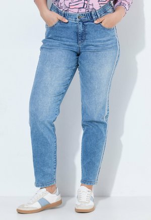 Person iført lyseblå denimjeans med hænderne i lommerne og hvide sneakers med blå striber står op ad en hvid væg.