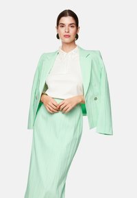 Mintgrüner Blazer mit strukturierten Schultern, kombiniert mit einem mintgrünen, plissierten Rock. Das Oberteil hat einen weißen Kragen mit Verzierungen.