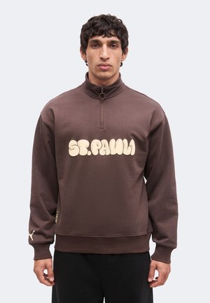 Mann trägt einen braunen Quarter-Zip-Sweatshirt mit "ST. PAULI" in großen cremefarbenen Buchstaben auf der Brust.