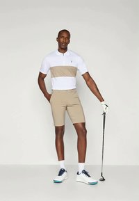 Lyhythihainen valkoinen ja beige kauluksellinen golfpaita, yhdistetty beigeihin shortseihin. Malli seisoo golfmailan kanssa, valkoiset kengät ja hansikkaat päällään.