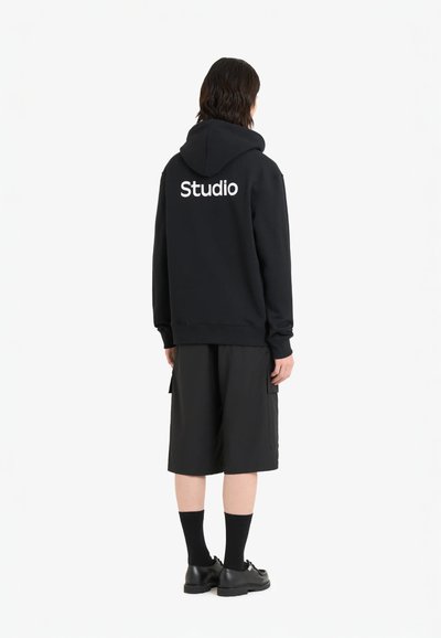 Haut à capuche noir avec "Studio" imprimé en blanc au dos, accompagné de shorts noirs. Présente une coupe décontractée et des poignets côtelés.