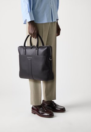 Black leather tote bag with dual handles, zip closure, minimalistic design, subtle branding, and structured shape, held by a person.