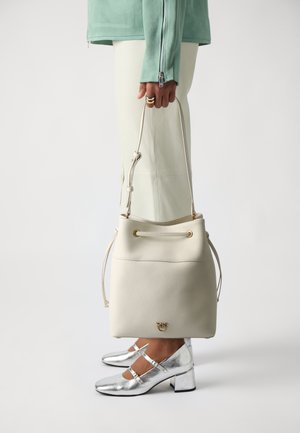 White leather bucket bag with drawstring closure and gold-tone hardware. Smooth texture, cylindrical shape. Worn alongside silver heels.