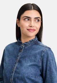 Jeansowa koszula w ciemnoniebieskim kolorze z falbaniastym kołnierzykiem, zapięciem na guziki, oraz kieszonką naszytą po lewej stronie. Gładka struktura i klasyczny design.