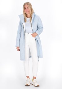 Donna che indossa un lungo cappotto azzurro chiaro con zip, pantaloni bianchi a vita alta, camicia a righe e sneakers bianche, in piedi davanti a uno sfondo bianco.