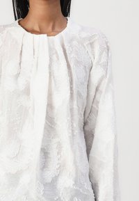 Hvit brodert bluse med rynket utringning, med intrikate blomstermønstre og lange ermer, laget av lett teksturert stoff.