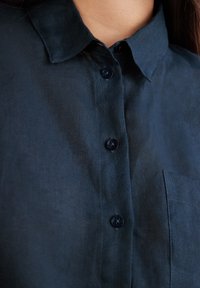 Camicia di lino blu navy con colletto, caratterizzata da un design con bottoni, due bottoni scuri e una tasca sul petto sul lato sinistro.
