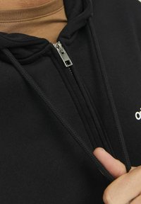 Zwarte zip-up hoodie van zachte stof met een zilveren rits en trekkoorden, voorzien van een klein wit logo aan de linkerkant.