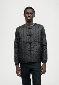 Giacca nera trapuntata con scollatura tonda, dotata di fibbie e orlo leggermente accorciato. Texture liscia e maniche lunghe con polsini arricciati.