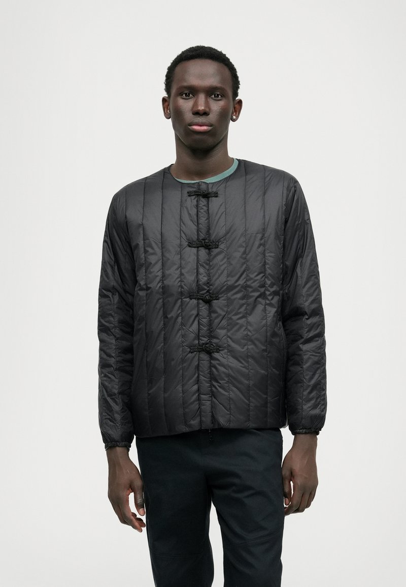 Giacca nera trapuntata con scollatura tonda, dotata di fibbie e orlo leggermente accorciato. Texture liscia e maniche lunghe con polsini arricciati.