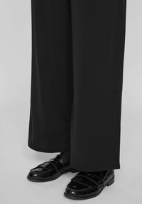 Crne hlače širokih nogavica s glatkom teksturom, uparene s sjajnim crnim mokasinkama s detaljnim šavovima i malim zlatnim akcentom.