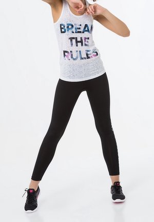 Camiseta sin mangas blanca con texto multicolor "BREAK THE RULES", combinada con mallas negras y zapatillas deportivas negras con acentos rosas.
