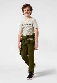 Šviesiai gelsvos spalvos marškinėliai su tamsiai mėlynu „Champion“ logotipu, derinami su alyvuogių žaliais sportiniais kelnėmis ir juodais sportbačiais su raudonomis detalėmis.