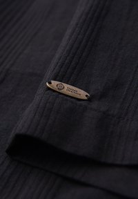 Étiquette métallique marron avec le texte "Ombre Heritage" attachée à un tissu noir texturé avec des motifs côtelés.