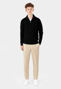Svart ribbet strikkegenser med glidelås i kragen, kombinert med beige skreddersydde bukser og hvite joggesko. Modellen står mot en lys bakgrunn.