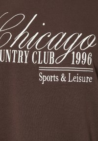Ruskea kangast-paita, jossa on valkoista brodeerattua tekstiä: "Chicago Country Club 1996" ja "Sports & Leisure" kauniilla kursiivilla.