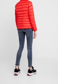 Rød polstret jakke med høj krave, ribbede manchetter og vandret quiltet design, parret med mørkeblå skinny jeans og sorte sneakers.