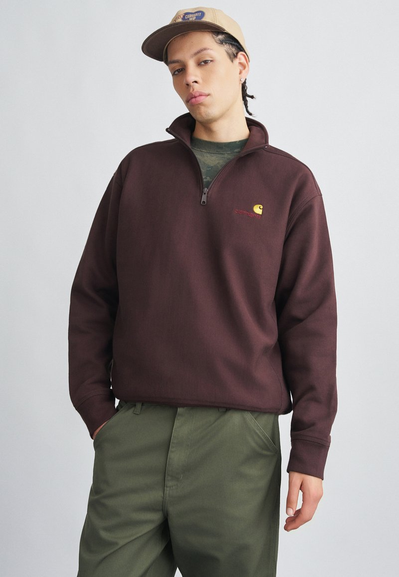Sweat-shirt en polaire bordeaux avec fermeture éclair partielle, arborant un petit logo jaune et rouge sur la poitrine et des poignets côtelés. Associé à un pantalon vert.