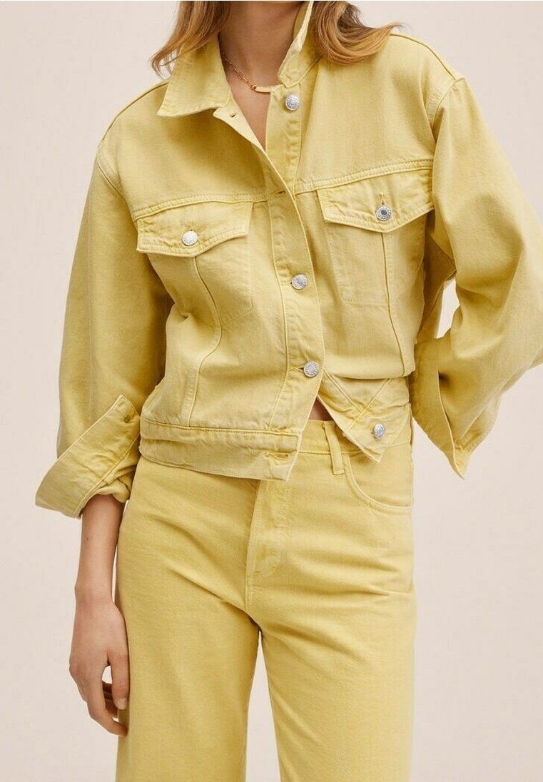 Femme portant une veste en jean jaune clair avec des boutons argentés et un jean jaune taille haute assorti sur un fond neutre.