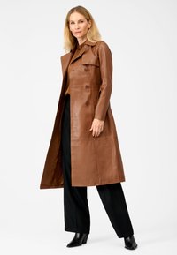 Brown leather long coat with a double-breasted design, notched collar, and side pockets. Smooth texture; paired with black trousers and boots.